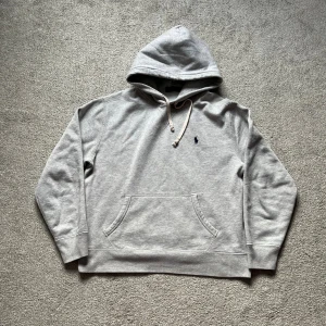 Grå hoodie från Polo Ralph Lauren - Snygg grå hoodie från Polo Ralph Lauren med klassisk broderad logga på bröstet. Tröjan har huva med snörning, känguruficka och långa ärmar. Perfekt för chill dagar och enkel att matcha med jeans eller joggers.