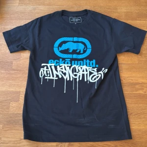 Svart Ecko Unltd t-shirt med blått tryck - Svart t-shirt från Ecko Unltd. med fet blått rhino-logga och graffiti-inspirerat vitt tryck på bröstet. Klassisk rund hals och korta ärmar. Materialet är 100% bomull, vilket gör den skön och lätt att bära. Perfekt för dig som gillar streetwear och coola prints.