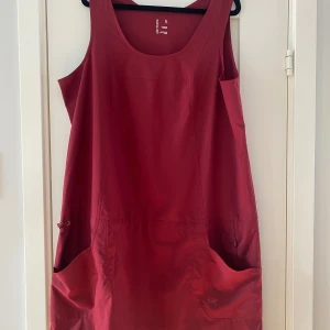 Röd ärmlös klänning från Arc'teryx - Snygg röd klänning från Arc'teryx med ärmlös design och rund halsringning. Klänningen har stora fickor framtill och en diskret snörning i midjan. Materialet är lätt och känns sportigt, perfekt för varma dagar. Loggan är tryckt på fickan för en cool touch. Mått Över bröstet: ca 59 cm Över höften: ca 61 cm Längd från axeln och ner: ca 98 cm  100% polyester   Kommer från ett djur och rökfritt hem