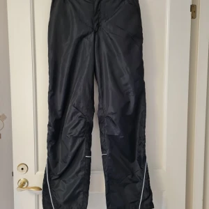 Svarta täckbyxor Seppälä Boys strl 164 - Svarta täckbyxor från Seppälä Boys i storlek 164. Byxorna har en loose passform, elastisk midja och är tillverkade i 100% polyester. Reflekterande detaljer vid benen.