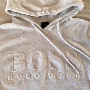 Hugo boss hoodie i frotté  - Säljer en vit hoodie från Hugo Boss med stor broderad logga på bröstet. Tröjan har huva med snörning, ribbade muddar och är tillverkad i mjuk frotté. Perfekt för dig som gillar stilrena och bekväma plagg med exklusiv känsla. Tröjan är i storlek M, herr-modell men passar även för dam. 