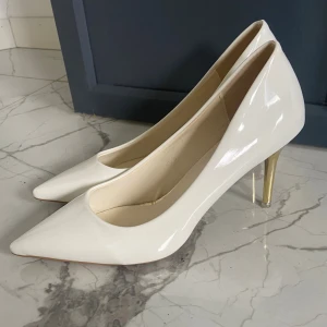 Vita pumps med guldklack - Snygga vita pumps med spetsig tå och glansig finish. Klacken är smal och i guld, vilket ger en lyxig touch. Skorna har en klassisk siluett och är tillverkade i syntetmaterial. Perfekta för dig som vill sticka ut med stilrena detaljer. Storlek 39