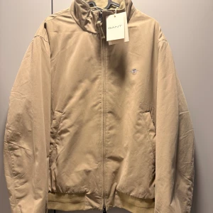 Beige vadderad jacka från GANT - Snygg beige vadderad bomberjacka från GANT med klassisk ståkrage och dragkedja framtill. Jackan har ribbade muddar vid ärmslut och nederkant, samt två sidofickor. Diskret GANT-logga broderad på bröstet. Perfekt för dig som gillar stilren design. Helt ny och oanvänd med prislapp kvar.