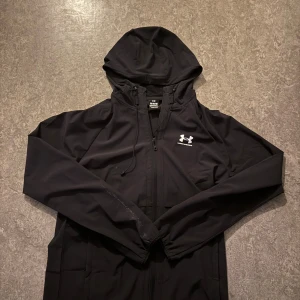 Svart vindjacka från Under Armour  - Svart vindjacka från Under Armour med huva och dragkedja framtill. Jackan har en stilren look med logga på bröstet och justerbar dragsko vid huvan. Tillverkad i lätt och smidig polyester, perfekt för blåsiga dagar.