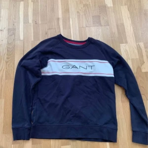 Mörkblå sweatshirt från GANT - Säljer en mörkblå sweatshirt från GANT med vit och röd rand över bröstet och stor GANT-logga. Tröjan har rund halsringning och långa ärmar. Perfekt för dig som gillar klassisk och stilren streetwear.