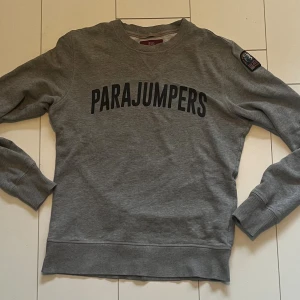 Grå fleece-tröja Parajumpers S - Snygg grå sweatshirt från Parajumpers i fleece-material med svart tryck över bröstet och ikonisk patch på ärmen. Tröjan har rund hals, ribbade muddar och en klassisk passform. Perfekt för dig som gillar stilrena och bekväma plagg.