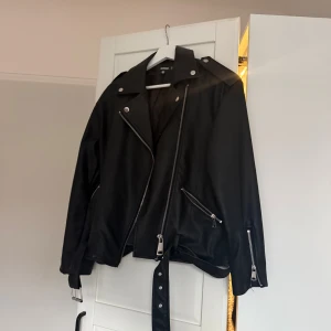 Svart skinnjacka från Missguided - Säljer en svart skinnjacka från Missguided i klassisk bikerstil med silvriga dragkedjor, knappar och bälte i midjan. Jackan har långa ärmar och coola detaljer som axelklaffar och snedställda fickor med dragkedja. Jackan är inte använd på några år men användes mycket då. 