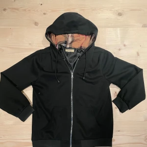 Svart hoodie från Burberry med dragkedja - Svart hoodie från Burberry med klassiskt rutigt foder i huvan. Tröjan har hel dragkedja framtill, fickor och ribbade muddar. Snygg och stilren design med exklusiv känsla, perfekt för dig som gillar premiumkänsla. Storlek M 