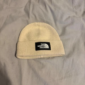 Beige mössa från The North Face - Säljer en stilren beige mössa från The North Face med ribbstickad design och uppvikt kant. Mössan har en svart patch med logga framtill och är tillverkad i 100% bomull, vilket gör den både mjuk och bekväm. Perfekt accessoar för kyliga dagar.