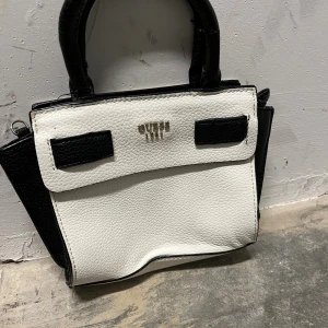 Vit och svart handväska från Guess - Snygg handväska från Guess i vit och svart skinnimitation med stilrena detaljer. Väskan har två handtag, dragkedja upptill och logotyp i metall framtill. Insidan är fodrad med mönstrat tyg och har en innerficka med dragkedja. Perfekt storlek för det viktigaste .Obs!-Något sliten på handtagen, syns på bild 3, annars bra skick.