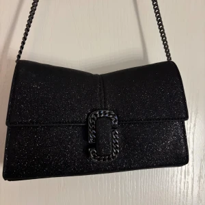 Svart glittrig axelväska Marc Jacobs - Snygg svart axelväska från Marc Jacobs med glittrigt material och kedjerem. Väskan har ett stilrent spänne framtill med kedjedetalj och praktiskt innerfack med dragkedja. Perfekt storlek för det viktigaste och en riktigt cool look. Nypris 3999