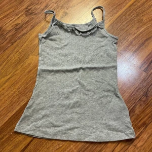 Grå ribbad linnetopp med spets från Esmara - Supersöt grå linnetopp från Esmara i ribbad bomull med smala axelband och spetsdetalj vid halsringningen. Perfekt för dig som gillar enkel stil med en liten twist. Sitter snyggt och är lätt att matcha med jeans eller kjol.