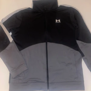 Svart och grå vindjacka Under Armour - Under Armour hoodie i svart och grått med hög krage och hel dragkedja framtill. Jackan har långärmade ärmar, två sidofickor och logga på bröstet. Materialet är mjukt och syntetiskt, perfekt för sportiga dagar eller chill. Snygg, clean design med normal passform.