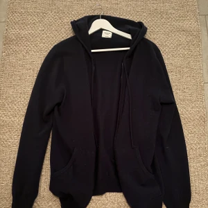 Mörkblå kashmir hoodie från John Henric (litet hål)  - Mysig mörkblå hoodie från John Henric i lyxig kashmir. Tröjan har huva, dragsko och två fickor framtill. Perfekt för dig som gillar stilrena och bekväma plagg med premiumkänsla. Har ett hål i ena armen som går att laga! 