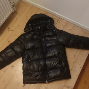 Svart pufferjacka Polo Ralph Lauren S - Svart pufferjacka från Polo Ralph Lauren med glansig finish, huva och dragkedja. Klassisk röd broderad logga på bröstet och RL-detalj på dragkedjan. Jackan är vadderad och har två fickor med dragkedja framtill. Perfekt för kalla dagar.