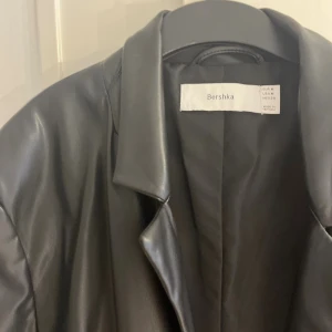 Svart skinnkavaj från Bershka - Snygg svart kavaj i skinnimitation från Bershka. Klassisk modell med krage, två framfickor med lock och en bröstficka. Rak passform och helfodrad insida, perfekt för en stilren look.