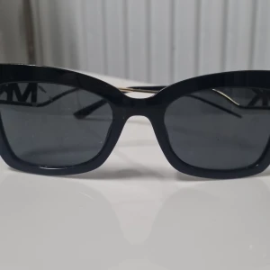 Svarta Michael Kors solglasögon - Snygga svarta solglasögon från Michael Kors med rektangulär form och mörka linser. Skalmarna har en cool detalj i guld med MK-logga i metall, vilket ger en lyxig vibe. Perfekt accessoar för att höja din stil på stan.