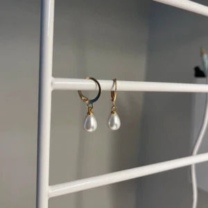 Guldiga örhängen med pärldroppar - Säljer ett par eleganta örhängen med guldfärgade ringar och droppformade vita pärlor. Perfekta för att ge en classy touch till din outfit. Enkla att matcha och riktigt stilrena. I rostfritt stål och handgjort 