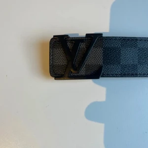 L&V bälte svart läder - Snyggt L&V bälte i klassiskt Damier Graphite-mönster med svart L&V-spänne. Bältet är gjort i skinn med rutigt mönster i mörkgrå och svarta toner. Insidan är ljusbrun och har präglad L&V-logga. Perfekt accessoar för att lyfta din outfit.