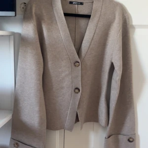 Beige kofta från Gina Tricot XS - Snygg beige kofta från Gina Tricot i mjukt material. Koftan har v-ringning, stora knappar framtill och dekorativa knappar vid ärmsluten. Perfekt för lager-på-lager och enkel att styla till olika outfits.