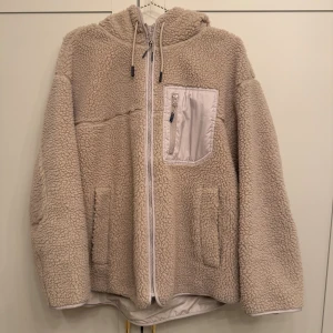 Beige teddyjacka med huva och dragkedja - Mysig beige teddyjacka från H&M med huva och hel dragkedja framtill. Jackan har en bröstficka med dragkedja och två sidofickor. Fodrad insida och snörning i huvan.