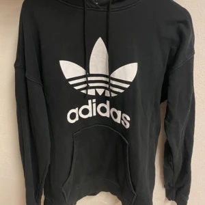 Svart Adidas hoodie med stor logga - Svart hoodie från Adidas med stor vit trefoil-logga och text på bröstet. Klassisk känguruficka framtill, huva med snörning och långa ärmar. Perfekt för en chill och sportig stil.