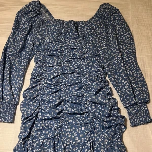 Blå blommig kortklänning från H&M - Supersöt blå kortklänning med vita småblommor, långa puffärmar och smock vid ärmslut. Klänningen har rynkade detaljer framtill som ger en snygg form och en fyrkantig ringning. Perfekt för dig som gillar romantisk och trendig stil. Klänningen är i storlek S