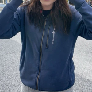 Blå kofta Sail Racing - Blå kofta från Sail Racing med dragkedja framtill och tryckt logga på bröstet och ryggen . Jackan har en sportig look och är perfekt för blåsiga dagar. Långärmad och med normal passform.