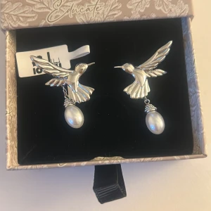 Ioaku silverfärgade kolibriörhängen med pärlor - Unika örhängen i form av silverfärgade kolibrier med hängande vita pärlor. Fågelns vingar är detaljerade och örhängena ger en cool och elegant vibe. Perfekta för dig som vill sticka ut med ett smycke som känns både lekfullt och stilrent.