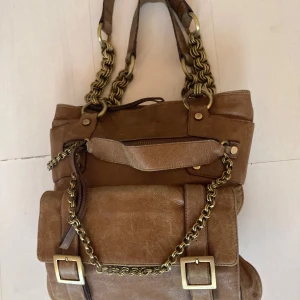 Brun handväska med kedjedetaljer - Snygg brun handväska i skinn från Karen Millen med grova guldfärgade kedjor och spännen. Väskan har flera fack med dragkedja som ger en unik look. Perfekt storlek för att få plats med allt du behöver och passar till många olika stilar.