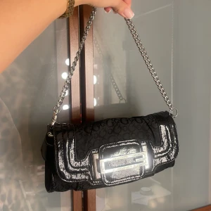 Svart axelväska från Guess med kedja - Snygg svart axelväska från Guess med glansiga detaljer och metallkedja som axelrem. Väskan har ett GG-mönster över hela ytan och en stor metallspänne framtill med Guess-logga. Perfekt storlek för det viktigaste och ger en cool vibe till din outfit. Går även att ta bort kedjan och ha som clutch 