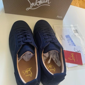 Mörkblå sneakers från Christian Louboutin - Exklusiva mörkblå sneakers från Christian Louboutin i mocka med ikonisk röd sula. Skorna har snörning, rund tå och diskret broderad logga på sidan. Insidan är ljusbeige och sulan är i svart och rött. Perfekta för dig som vill sticka ut med lyxig streetstyle.
