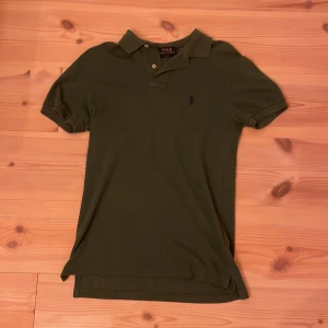 Olivgrön pikétröja från Ralph Lauren - Snygg olivgrön pikétröja från Ralph Lauren med klassisk krage och två knappar. Tröjan har korta ärmar och den ikoniska broderade loggan på bröstet. Perfekt för en clean och stilren look.