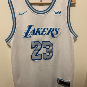 Lakers LeBron James #23 baskettröja - Säljer en vit baskettröja från Nike med Lakers-logga och LeBron James namn och nummer 23 i blått och silver. Tröjan är ärmlös med blå och silverfärgade detaljer runt hals och ärmhål. Materialet är mesh för extra ventilation och sportig känsla.