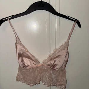 Linne - Säljer en champagnefärgad topp/bh med tunna axelband och justerbar rygg. Toppen har v-ringad form, ärmlös design och är gjord i glansig satin med romantiska spetsdetaljer längs kanterna och nederdelen. 