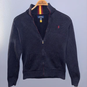 Mörkblå ziptröja från Polo Ralph Lauren - Mörkblå stickad ziptröja från Polo Ralph Lauren med röd broderad logga på bröstet. Tröjan har hög krage, dragkedja framtill och två fickor. Ribbstickade muddar vid ärmslut och nederkant. 