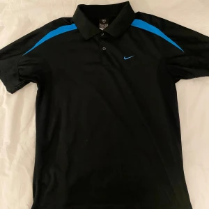 Svart och blå Nike pikétröja - Snygg svart pikétröja från Nike med blå detaljer på axlarna och klassisk krage med två knappar. Tröjan har korta ärmar och är tillverkad i ett lätt och sportigt material som passar perfekt för träning eller en chill look.