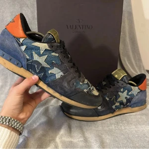 Valentino Rockrunner sneakers stjärnmönster - Unika Valentino Rockrunner sneakers med blått och svart stjärnmönster i denim och mocka. Orange häl med nitar i stjärnform och blå detaljer på sulan. Svarta skosnören och guldiga logodetaljer på plösen. Perfekt för dig som vill sticka ut med lyxig streetstyle.
