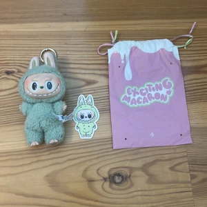 Exciting Macaron grön labubu - Söt grön shoppingväska med dragsko och trycket 'Exciting Macaron' i pastellfärger. Väskan har vita detaljer upptill och färgglada snören. Perfekt för småsaker eller som presentpåse, med lekfull design som sticker ut. Den är helt äkta och påse, kort följer med