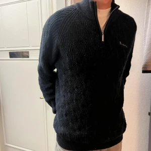  zip-tröja Bondelid - Mörk stickad tröja från bondelid med snyggt strukturmönster och half zip-dragkedja vid halsen. Tröjan har lång ärm och hög krage, perfekt för kyliga dagar. Broderad logga på bröstet ger en clean look.