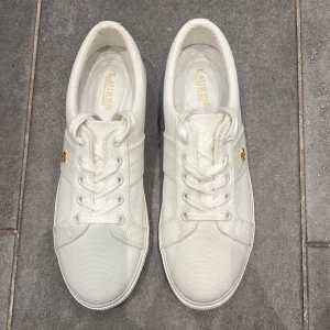 Vita sneakers från Lauren Ralph Lauren - Snygga vita sneakers från Lauren Ralph Lauren i canvas med klassisk rund tå och platt sula. Skorna har snörning och en liten guldfärgad RL-detalj på sidan som ger en exklusiv touch. Perfekta för en clean och fräsch look.