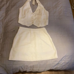 Beige/vit omlottväst med knyt,kjol - Snygg beige/vit  väst med omlott och knyt i nacken med tillhörande kort kjol  med resor på baksidan. Använd 1 gång. Mycket bra skick.Västen har v-ringning, knappar framtill och två dekorativa fickor. Tillverkad i ett linneliknande material som känns luftigt och fräscht. Perfekt att matcha med kjol eller jeans för en clean look.vet inte om det är linne material men mjuk och skön.