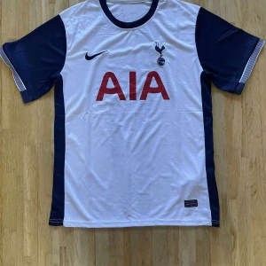Tottenham Hotspur Nike fotbollströja - Säljer en Tottenham Hotspur matchtröja från Nike. Tröjan är vit med mörkblå ärmar och detaljer, röd AIA-logga på bröstet och klubbmärke på vänster sida. Tillverkad i lätt och ventilerande material, perfekt för fotboll eller supporterliv.