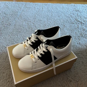 Michael Kors vita och svarta sneakers - Snygga sneakers från Michael Kors i vitt skinn med svart detalj på sidan och guldfärgade MK-loggor på snörningen. Klassisk rund tå och platt sula. Perfekta för dig som vill ha en stilren och trendig look med exklusiv känsla. Aldrig använda 