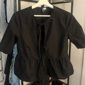 Svart blus med knyt från Concept - Svart blus från Concept med knyt framtill och volang nertill. Modellen har kort ärm och en luftig passform, perfekt för dig som gillar en enkel men ändå trendig look. Blusen har rund halsringning och är tillverkad i ett lätt bomullstyg.