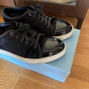  Lanvin med gläns  - Snygga svarta sneakers från Lanvin med vit sula och snörning. Skorna har en blank tå i skinn och resten av ovandelen är i mocka, vilket ger en stilren och klassisk stil . Hör av er vid frågor! Box och dustbag även äkthetsbevis kvitto följer med !