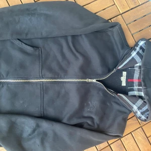 Svart hoodiejacka från Burberry M - Svart hoodiejacka från Burberry med klassiskt rutigt foder i huvan och dragkedja framtill. Jackan har broderad logga på bröstet, två fickor och ribbade muddar. Materialet är mjukt och bekvämt, perfekt för en chill och stilren look. Finns lite små fläckar som går bort genom tvätten