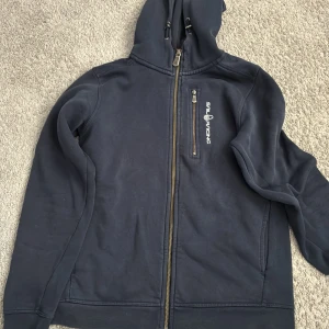 Mörkblå hoodie från Sail Racing - Mörkblå hoodie från Sail Racing med hel dragkedja, huva och en bröstficka med dragkedja. Snygg logga på bröstet och mjukt material som passar perfekt till kyliga dagar. Klassisk och sportig look med långa ärmar och ribbade muddar.