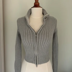 Grå ribbad kofta med dragkedja - Snygg grå ribbstickad kofta med hög krage och hel dragkedja framtill. Modellen är croppad och har långa ärmar, perfekt för lager-på-lager. Materialet är stickat och ger en skön och mjuk känsla. Passformen är slim och framhäver midjan.