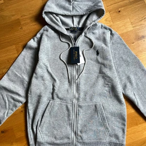 Grå hoodie från Polo Ralph Lauren - Snygg grå hoodie från Polo Ralph Lauren med dragkedja och huva. Slim fit-modell med mjukt bomullsmaterial och klassisk logga broderad på bröstet. Utrustad med snörning i huvan och två praktiska fickor framtill. Perfekt för chill dagar.